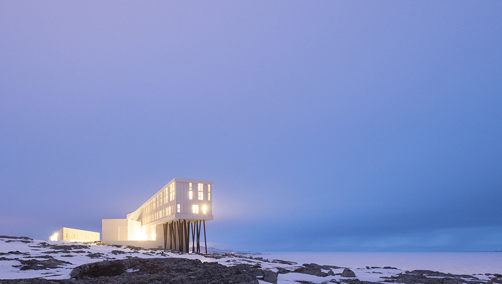 The image showcases the Fogo Island Inn, situated on a rugged rocky outcrop, its white façade glistening in the light. In the foreground, three buildings with flat roofs are nestled among snow-covered rocks, while the main building stands tall on stilts, featuring numerous windows and illuminated from within. The surrounding landscape is dotted with scattered patches of snow, set against a serene blue sky with a soft pink hue on the horizon.