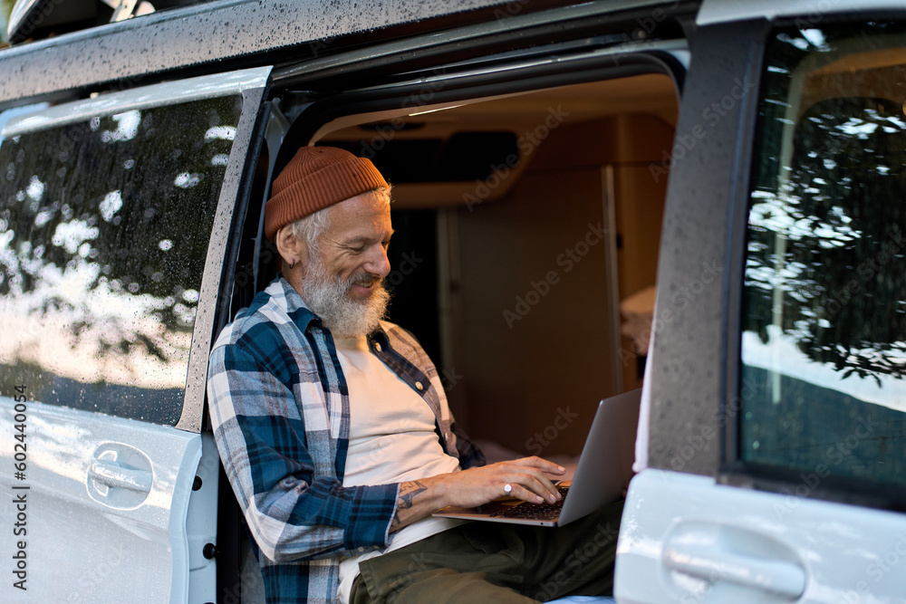 The image shows a man sitting in the back of a van, working on his laptop. * The man is wearing a brown beanie and a blue-and-white plaid shirt. * He has white hair and a beard. * His face is blurred out. * The van's interior is visible through the open door. * There are trees reflected in the windows. * The van appears to be parked near some trees or foliage. * The overall atmosphere suggests that the man is working remotely from his vehicle. The image conveys a sense of flexibility and productivity, as the man is able to work comfortably in a non-traditional workspace.