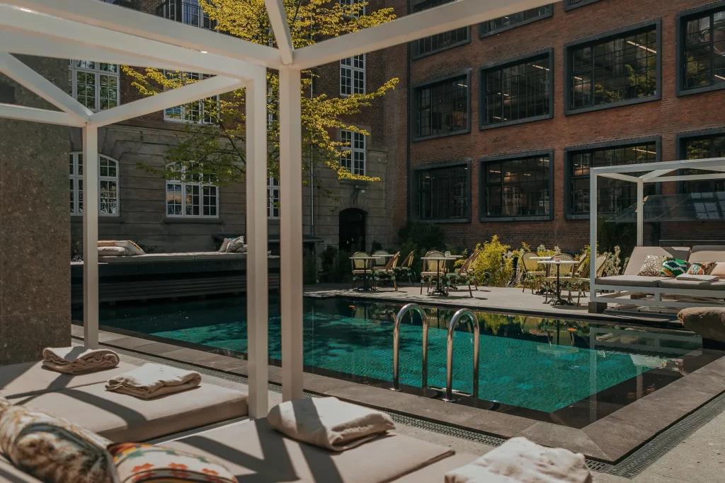 This image depicts a serene poolside setting, with two beige lounge chairs positioned beside the pool. The lounge chairs feature plush cushions adorned with white towels neatly folded on top. A white metal frame supports the canopy above, adding to the relaxing ambiance. The pool's crystal-clear water reflects the surrounding environment, while a few vibrant yellow flowers in the background add a pop of color to the scene. In the distance, a brick building with multiple windows and a brown exterior provides a subtle backdrop to the tranquil atmosphere. The overall setting exudes a sense of calmness and relaxation, inviting the viewer to unwind and soak up the serenity.