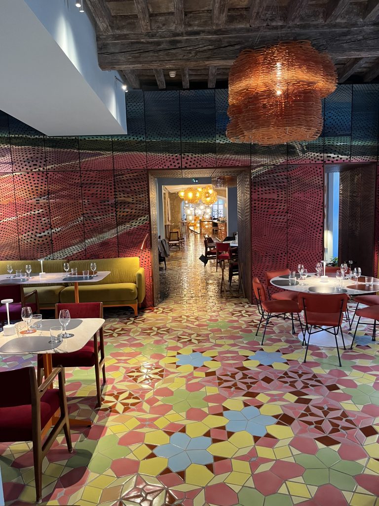 Alt tag: "Interior of a modern restaurant featuring colorful geometric tile flooring, stylish seating with yellow and red upholstery, and decorative lighting. The entrance leads to a warmly lit dining area."