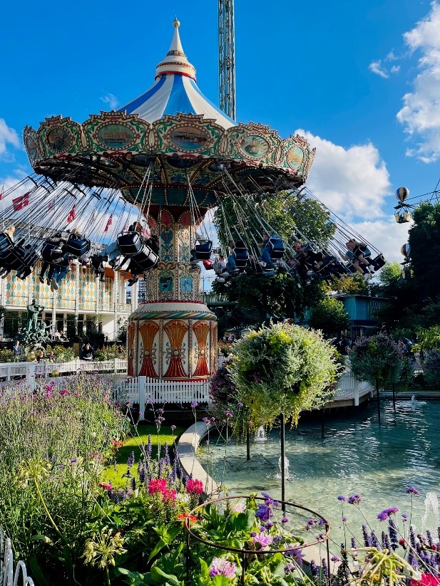 Alt tag: A colorful swing carousel in an amusement park surrounded by vibrant flowers and greenery, with a blue sky and fluffy clouds above.