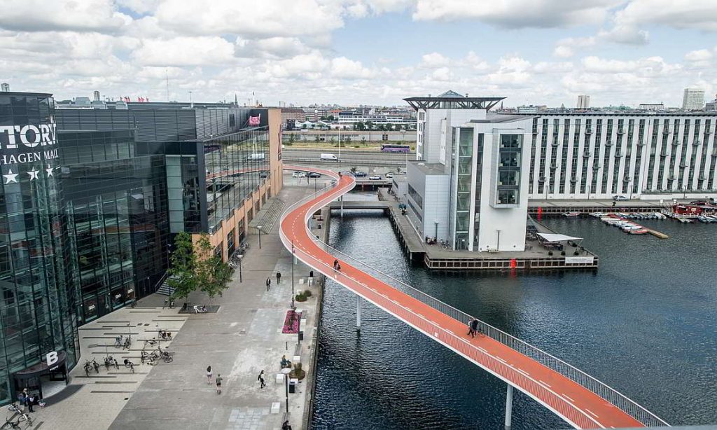 Aerial view of a modern urban landscape featuring a red pedestrian bridge over a waterway, connecting buildings and commercial spaces, with cloudy skies overhead.