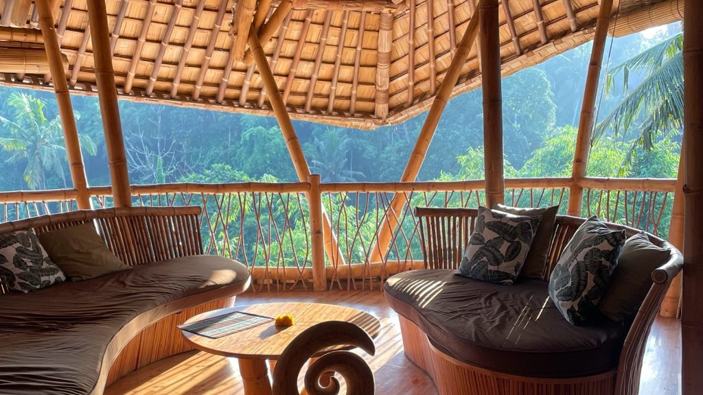 This image depicts the interior of a bamboo-walled room, featuring two circular benches and a small table situated in front of a large window offering a breathtaking view of the forest. The room's rounded walls are crafted from bamboo, complemented by light-colored wood flooring that harmonizes with the natural surroundings. The circular benches, adorned with dark brown cushions, are accompanied by throw pillows showcasing an abstract green design. A small table, made of the same material as the floor and wall, holds a few items on its surface. Beyond the window, a lush forest landscape unfolds, with trees and foliage visible in the distance. The overall ambiance exudes a sense of serenity and tranquility, evoking feelings of relaxation and calmness.