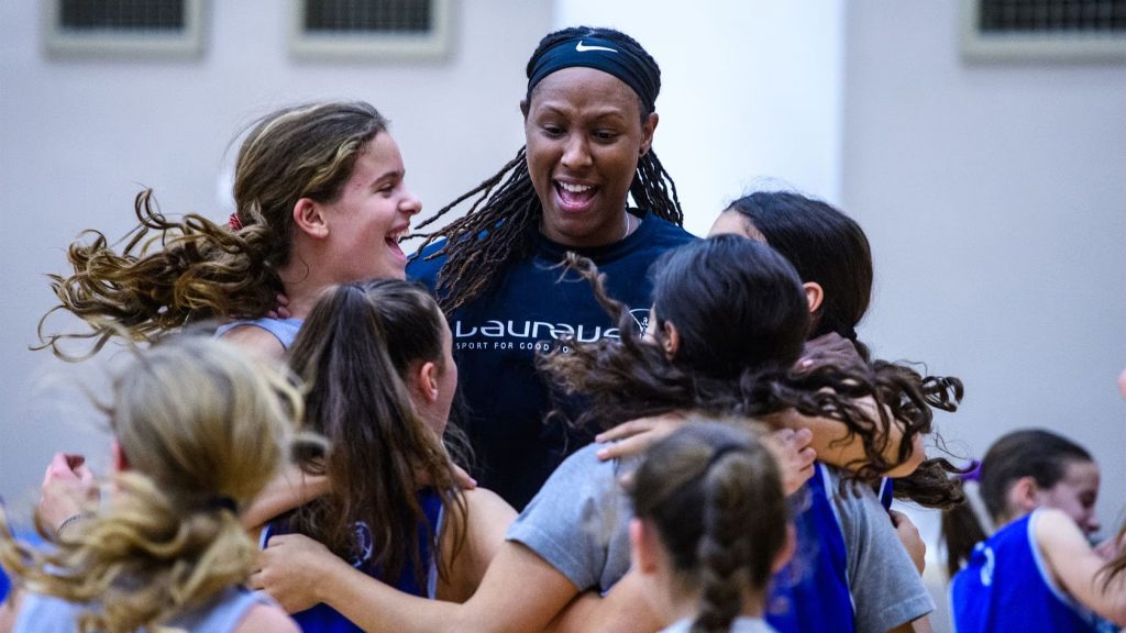 A group of young female basketball players joyfully celebrates with a smiling coach in a gymnasium, showcasing teamwork and enthusiasm.
