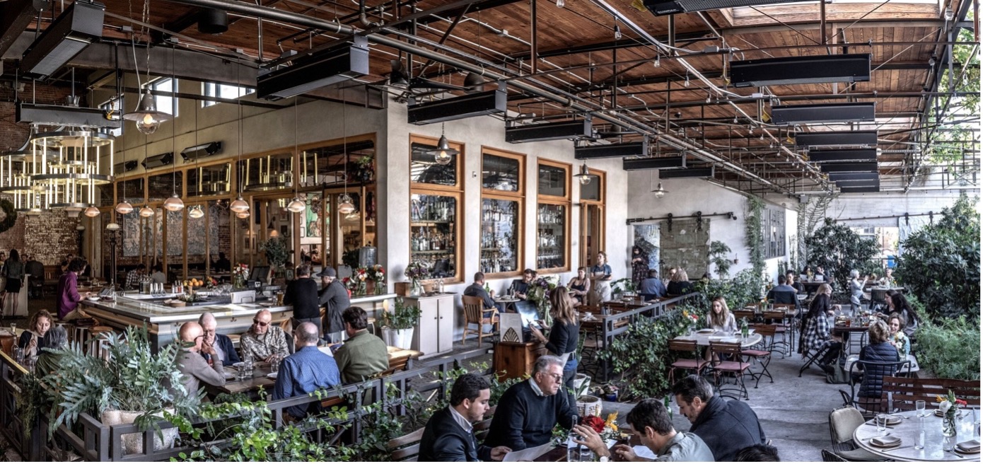 The image depicts the interior of a restaurant, with a long bar and tables filled with diners. In the foreground, a large white bar stretches across the room, featuring a row of stools along its length. Behind the bar, a wall of windows allows natural light to pour in, while a white countertop is adorned with various bottles and glassware. The background reveals more tables and chairs, where patrons are engaged in conversation or enjoying their meals. The atmosphere appears lively and bustling, with an overall ambiance that suggests a casual dining experience.
