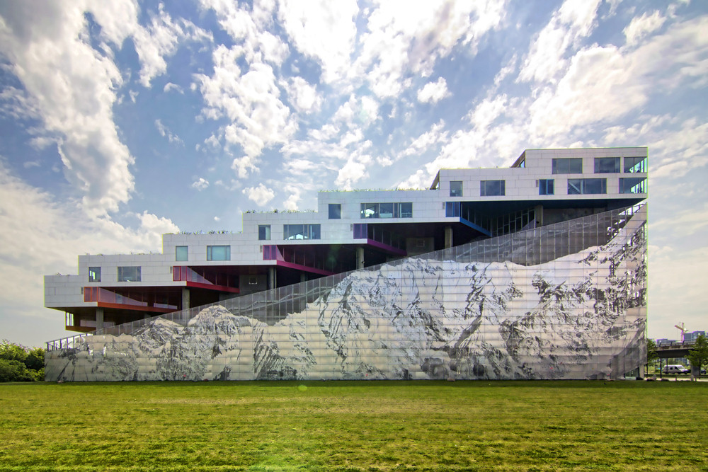 Alt tag: A modern, multi-level building featuring a striking facade that blends concrete and vibrant colored sections with a large, mural-like design resembling a mountain landscape, set against a bright sky with scattered clouds. Green grass is visible in the foreground.