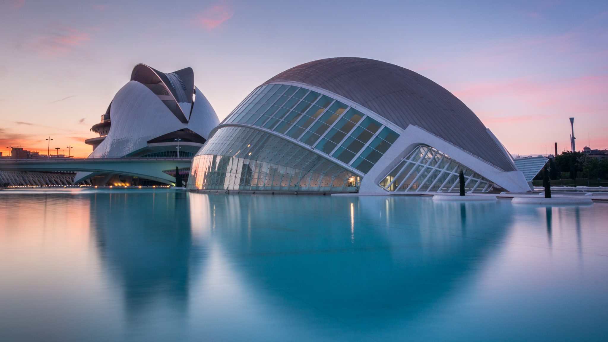 This photograph showcases the striking L'Hemisfèric building in Valencia, Spain, during sunset. The building's distinctive design features three interconnected spherical structures, comprising a planetarium and IMAX cinema on the left, an art gallery and cafe on the right, and a laserium and bar in the center. The curved glass walls of the middle sphere reflect vibrant hues, while its central opening frames the adjacent buildings. In the foreground, the blue water creates a stunning reflection of the building's curves. The background reveals additional structures in the distance, with the sky transitioning from orange to pink as it meets the horizon, indicating an early evening setting.