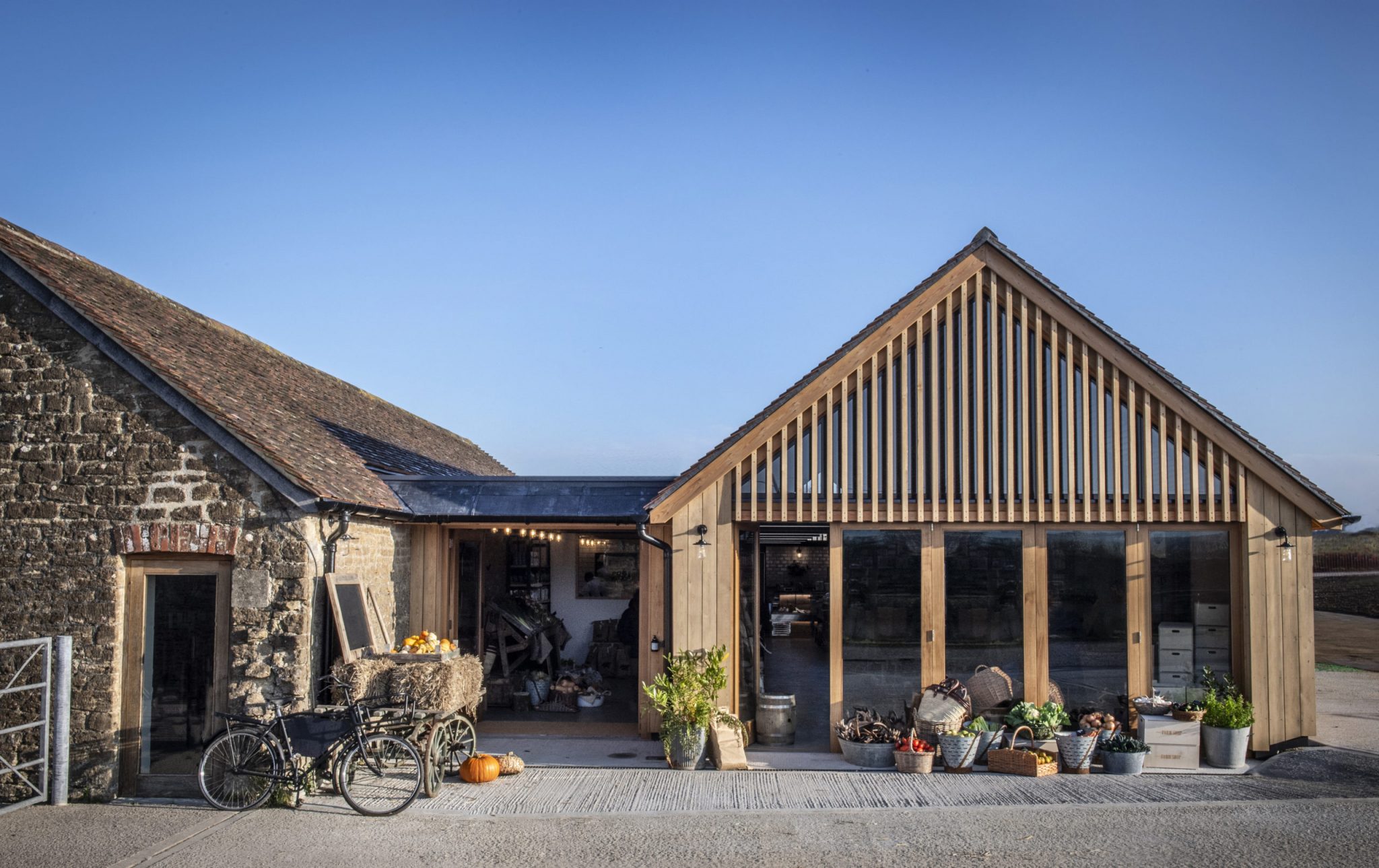 The image depicts the exterior of a rustic, stone-walled building with a brown roof, accompanied by an adjacent wooden structure featuring a pitched roof. In the foreground, a black bicycle is parked in front of the stone wall, adorned with pumpkins and gourds on its back carrier. A large burlap sack filled with hay sits beside the bike, while a few potted plants are scattered around the area. The stone building's entrance is situated to the left of the image. The wooden structure, located to the right, boasts a pitched roof and features several doors and windows. Various gardening tools and supplies are visible on the ground outside the building. In the background, a blue sky stretches out, providing a serene atmosphere.