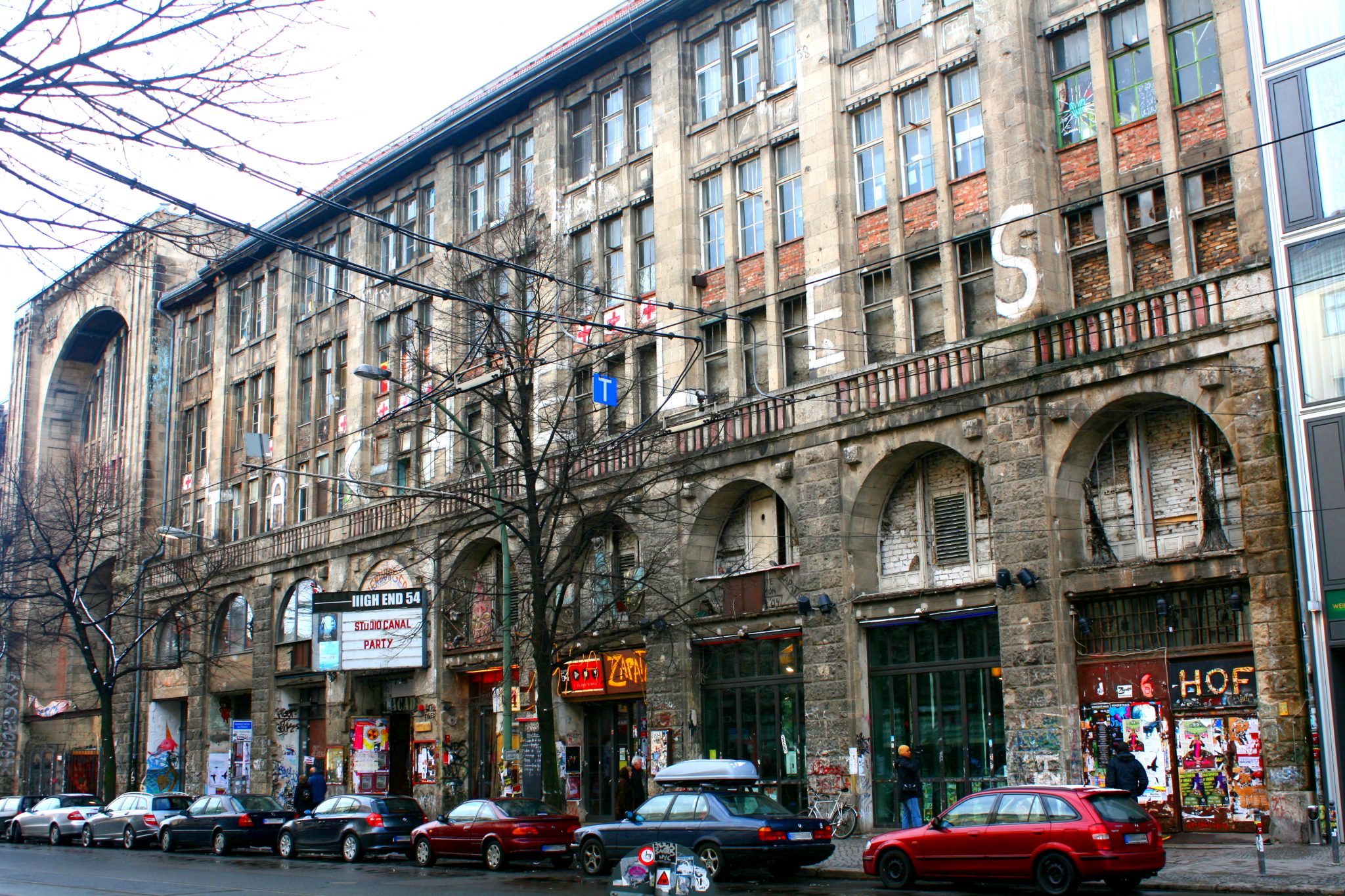 The image depicts a city street lined with buildings, showcasing a mix of old and new architecture. The scene is set against a cloudy sky. * **Buildings:** * The buildings are primarily made of stone or brick, with some having newer additions. * Some structures appear to be abandoned or in disrepair, while others have been renovated. * A few buildings feature ornate details such as arches and balconies. * **Street Scene:** * The street is relatively narrow, with a mix of parked cars and pedestrians. * Several shops and restaurants line the street, including a coffee shop with outdoor seating. * A large tree stands in front of one of the buildings, adding some greenery to the scene. * **Sky:** * The sky is overcast, casting a grayish hue over the entire image. * No visible sun or clouds are present, giving the impression of an overcast day. In summary, the image presents a charming urban street with a blend of old and new architecture, set against a gloomy yet atmospheric backdrop.