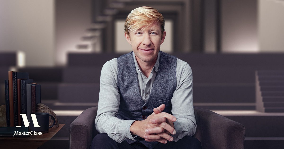 Man with blonde hair seated on a couch, wearing a gray vest, smiling confidently in a modern, softly lit environment with subtle background elements and a MasterClass logo in the corner.