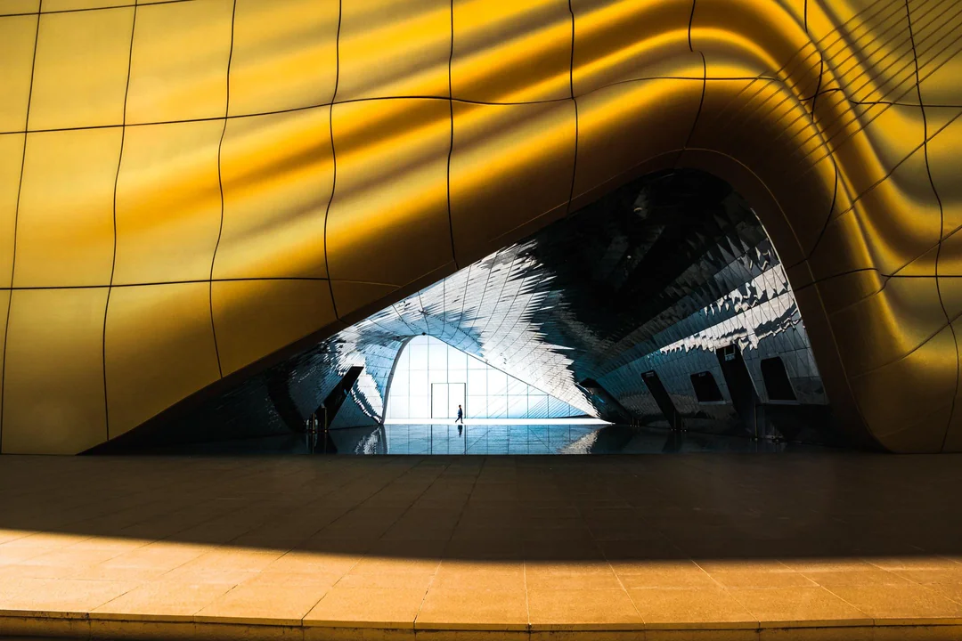 The image showcases the interior of a large, modern building featuring curved walls and a high ceiling. In the foreground, the yellow wall is composed of square panels that are slightly curved, with light reflecting off them. The wall curves to form an arched opening in the background, where a person can be seen standing at the far end, gazing out into the distance. The archway leads to another room or hallway beyond. The overall atmosphere suggests a contemporary architectural design, possibly a public building such as a train station or airport terminal.