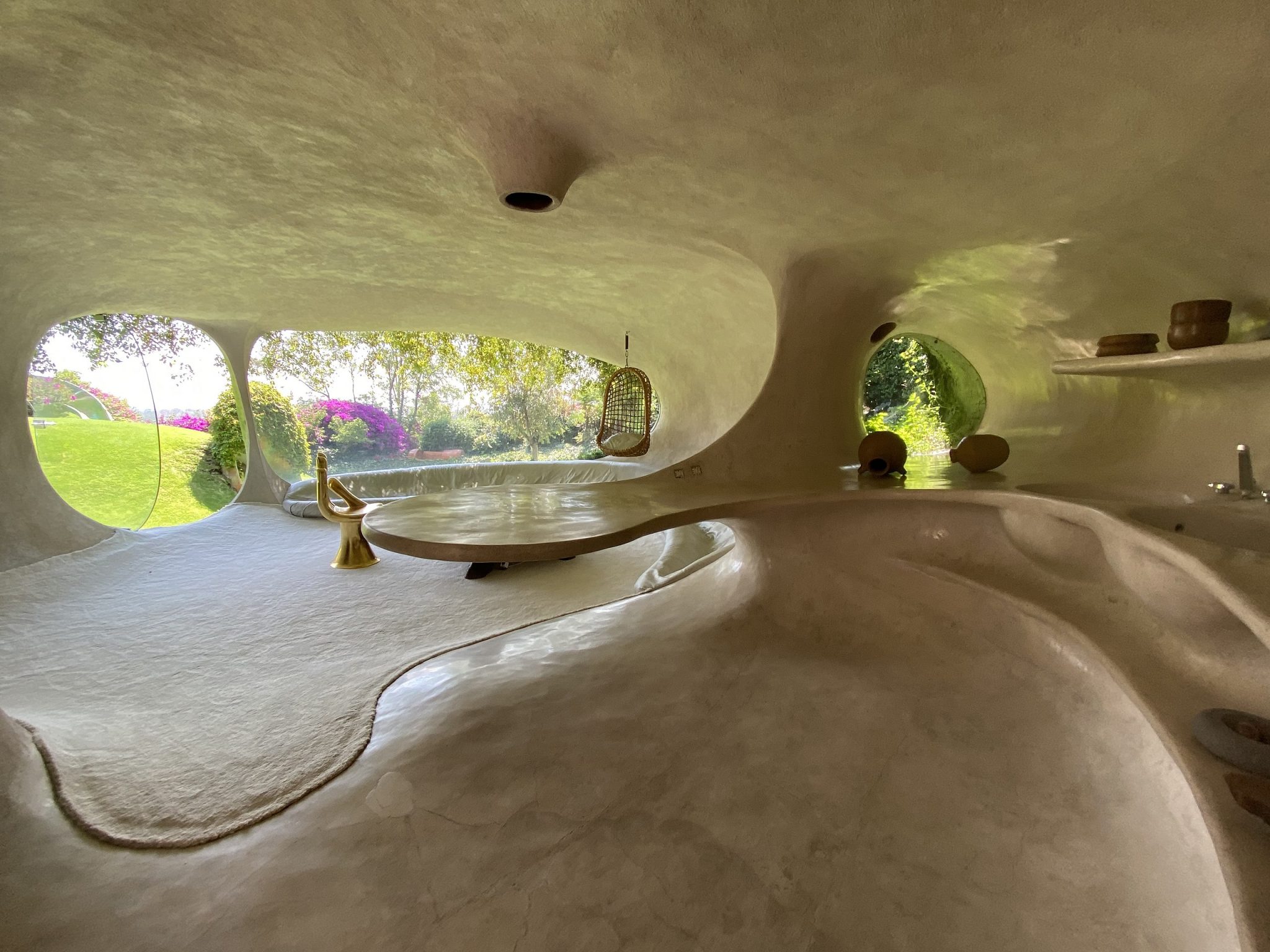 The image depicts the interior of a unique, organic-shaped room with an all-white color scheme. The space features rounded walls and ceiling, adorned with several circular windows that provide natural light. The floor is covered in white marble or stone, while various furniture pieces are scattered throughout the room. A long, curved bench runs along the right side wall, accompanied by a few shelves on the opposite wall. Two large, round tables sit in front of the windows, and several chairs are placed around the room. The overall atmosphere suggests that this is an interior design concept or a finished space designed for relaxation and contemplation. The use of organic shapes and natural materials creates a calming ambiance, perfect for unwinding or enjoying quiet moments.