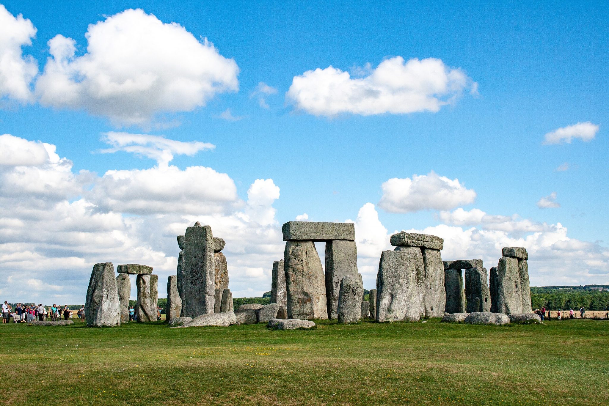 Alt tag: "Stonehenge, a prehistoric stone monument, stands prominently against a blue sky filled with fluffy white clouds, with visitors observed in the background on a grassy field."