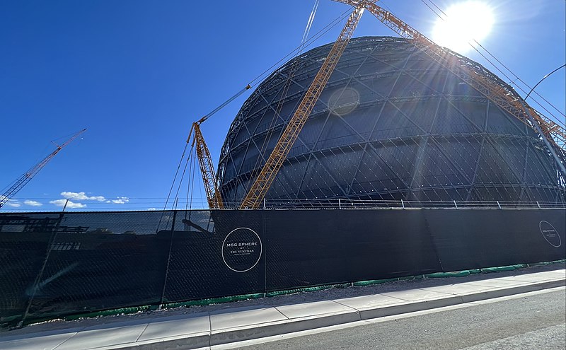 The image depicts the construction of a large, dome-shaped building under a clear blue sky with minimal clouds. * **Construction Site:** * A large, black metal structure resembling a dome is being constructed. * The site features two cranes positioned on either side of the dome, which are likely used for lifting and moving heavy materials during construction. * The building's framework consists of a series of interconnected steel beams that form the shape of a dome. * **Background:** * The sky above is a bright blue with only a few small clouds visible in the distance. * The overall atmosphere suggests a sunny day, possibly during the late morning or early afternoon. * **Signage and Markings:** * A large black banner stretches across the front of the construction site, featuring white text that reads "MSG SPHERE" within a circular logo. * Below this, smaller text is illegible due to its small size. In summary, the image captures the ongoing construction of a large dome-shaped building on a sunny day. The use of cranes and steel beams indicates that the structure is being built using heavy machinery and materials.