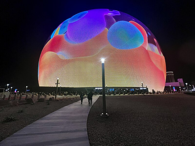 The image depicts the exterior of the MSG Sphere at The Venetian Resort, a large spherical structure with a vibrant, colorful design on its surface. * **Sphere:** * Large, spherical shape * Colorful design featuring various colors and patterns * Appears to be made of a translucent material, allowing light to pass through * **Lighting:** * Bright lights illuminating the sphere from within * Lights are likely used for decorative purposes or to highlight specific features of the structure * **Surroundings:** * Desert landscape surrounding the sphere * Sparse vegetation and rocky terrain visible in the background The image showcases the unique design and lighting features of the MSG Sphere at The Venetian Resort, highlighting its striking appearance against a desert backdrop.