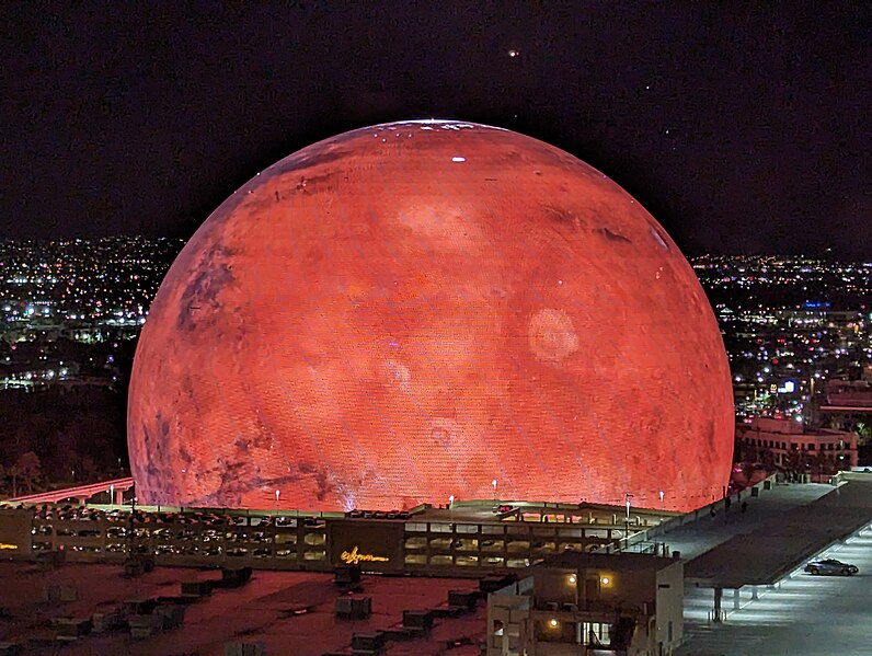 The image depicts the exterior of the Las Vegas Sphere, a large spherical structure with a distinctive red-orange hue, illuminated at night. In the foreground, the sphere is situated on a rooftop, surrounded by other buildings in the background. The surrounding area appears to be a cityscape, with various structures and lights visible against the dark sky. The overall atmosphere of the image suggests that it was taken at night, likely during an event or performance held within the sphere's interior.