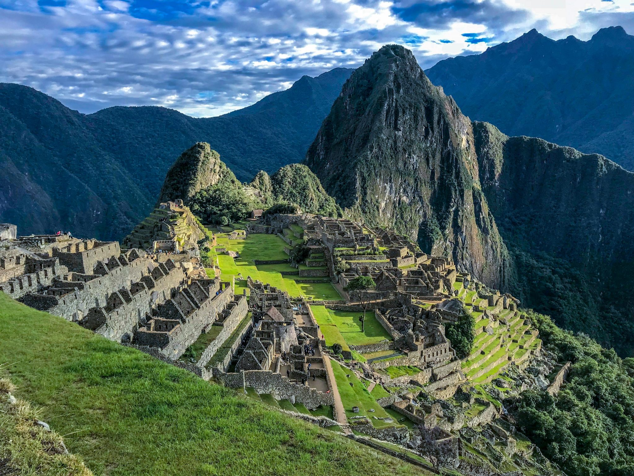 Alt tag: A panoramic view of Machu Picchu, an ancient Incan city, nestled in the Andes Mountains, featuring terraced structures and lush greenery under a dramatic sky.