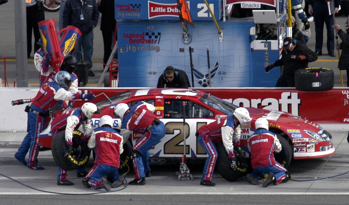 The image depicts a NASCAR pit crew in action, with their team's car being serviced during a race. * A red and white race car is on the ground: + The car has a distinctive design with red and white stripes running along its sides. + It appears to be a stock car, likely from the NASCAR series. * The car has a large number 21 on it: + The number 21 is prominently displayed in gold letters on the side of the car. + This suggests that the car may be driven by a driver with the number 21 as their racing number. * A group of people are working under the hood: + Several individuals, likely pit crew members, are gathered around the front of the car. + They appear to be engaged in various tasks, such as changing tires or refueling the vehicle. Overall, the image captures a critical moment in a NASCAR race, highlighting the importance of teamwork and quick thinking during pit stops.