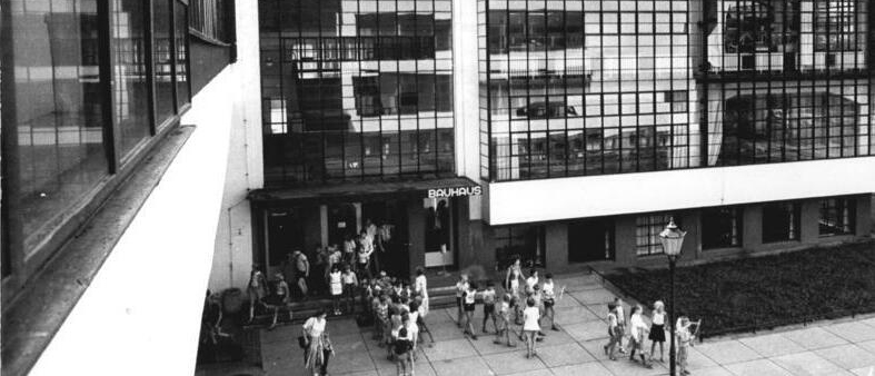 A historical black-and-white photo of the Bauhaus building, featuring a group of people walking towards the entrance, showcasing the building's modernist architecture with large windows and geometric lines.