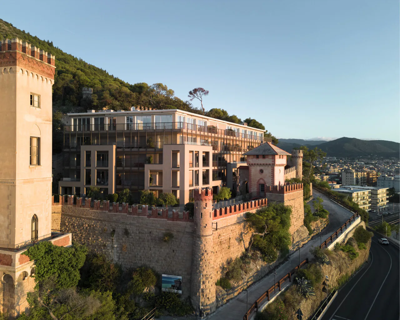 A modern building integrated with a historic castle, situated on a hillside with lush greenery, overlooking a city landscape and roadway below.