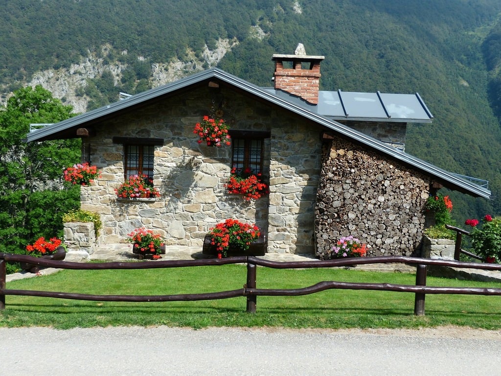 This image depicts the exterior of a charming stone house nestled in a mountainous region, showcasing its rustic charm. The one-story house features a light-colored stone facade with dark brown trim around windows and doors, complemented by a distinctive red brick chimney. A substantial woodpile is neatly stacked against the right side of the building. The roof is composed of dark gray metal panels, while several potted plants adorn the front yard. In the foreground, a split-rail fence stands out against a backdrop of lush green trees and mountains. The image presents a serene and picturesque scene, evoking a sense of tranquility and connection to nature.