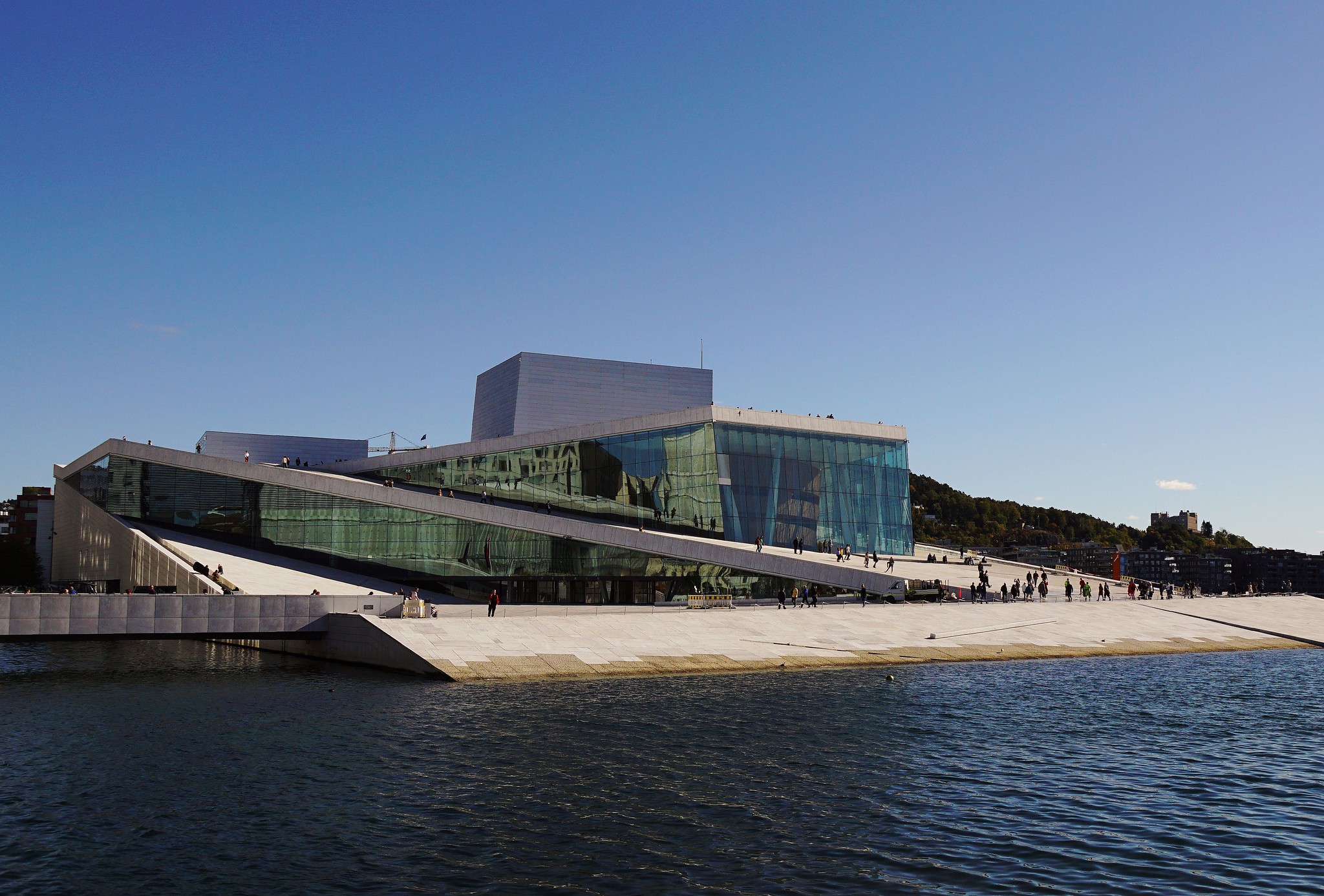 The image depicts the Oslo Opera House in Norway, showcasing its unique design and stunning surroundings. * The building: + Features a sleek, modern architecture with large glass windows. + Has a distinctive "wavy" shape that blends seamlessly into the surrounding landscape. + Appears to be made of concrete or stone, giving it a sturdy yet elegant appearance. * The water: + Surrounds the building on three sides, creating a sense of tranquility and connection to nature. + Reflects the building's design, adding depth and visual interest to the scene. * The sky: + Is clear and blue, providing a beautiful backdrop for the opera house. + Adds to the overall sense of serenity and grandeur. The image effectively captures the essence of the Oslo Opera House as a symbol of cultural excellence and architectural innovation.