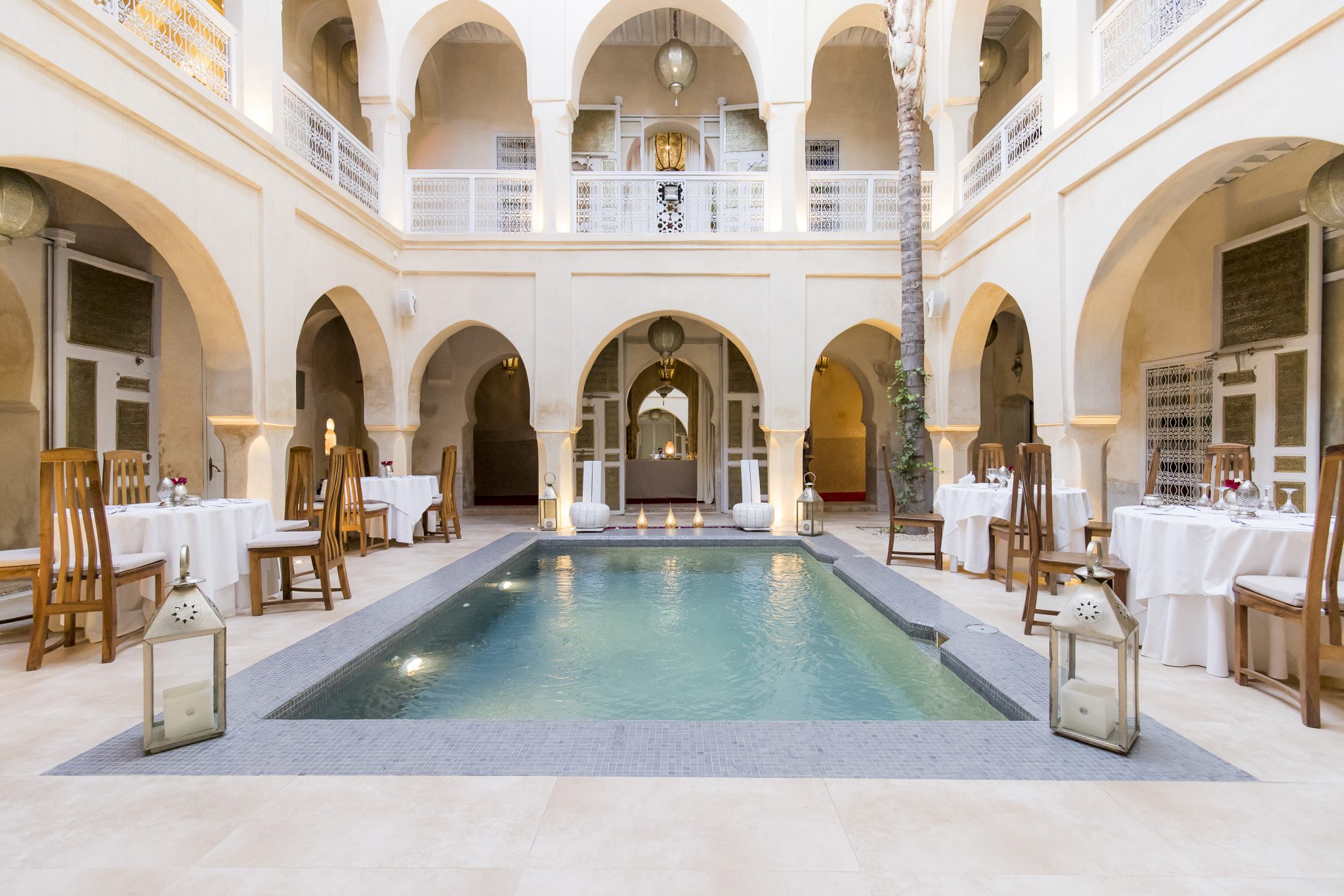 The image presents a serene courtyard with a pool, adorned with tables and chairs, set against the backdrop of an elegant building featuring arches. * **Pool** + Located in the center of the courtyard + Surrounded by tables and chairs on all sides + Rectangular shape with a gray tiled border + Water is calm and peaceful * **Building** + White walls and columns + Arches connecting the different levels + Balconies on the upper floors + Large windows allowing natural light to enter * **Furniture** + Tables covered with white tablecloths + Wooden chairs placed around each table + Lanterns or candles on some tables, adding a warm ambiance The image showcases a tranquil and inviting courtyard, perfect for relaxation or socializing. The combination of the pool, elegant building, and comfortable furniture creates a sense of serenity and sophistication.