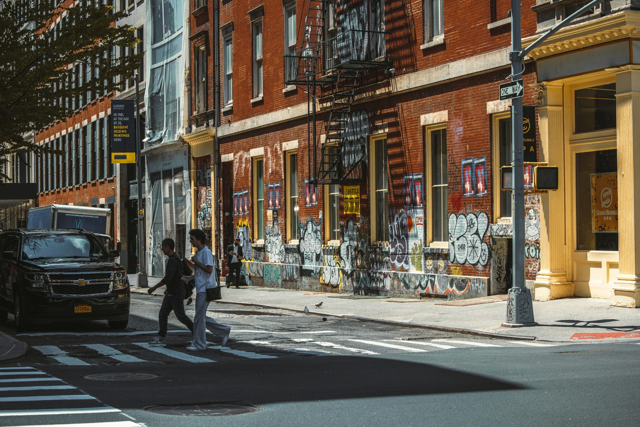 Urban street scene featuring pedestrians crossing a zebra-striped crosswalk in front of a brick building adorned with colorful graffiti, flanked by storefronts and a parked black SUV on a sunny day.