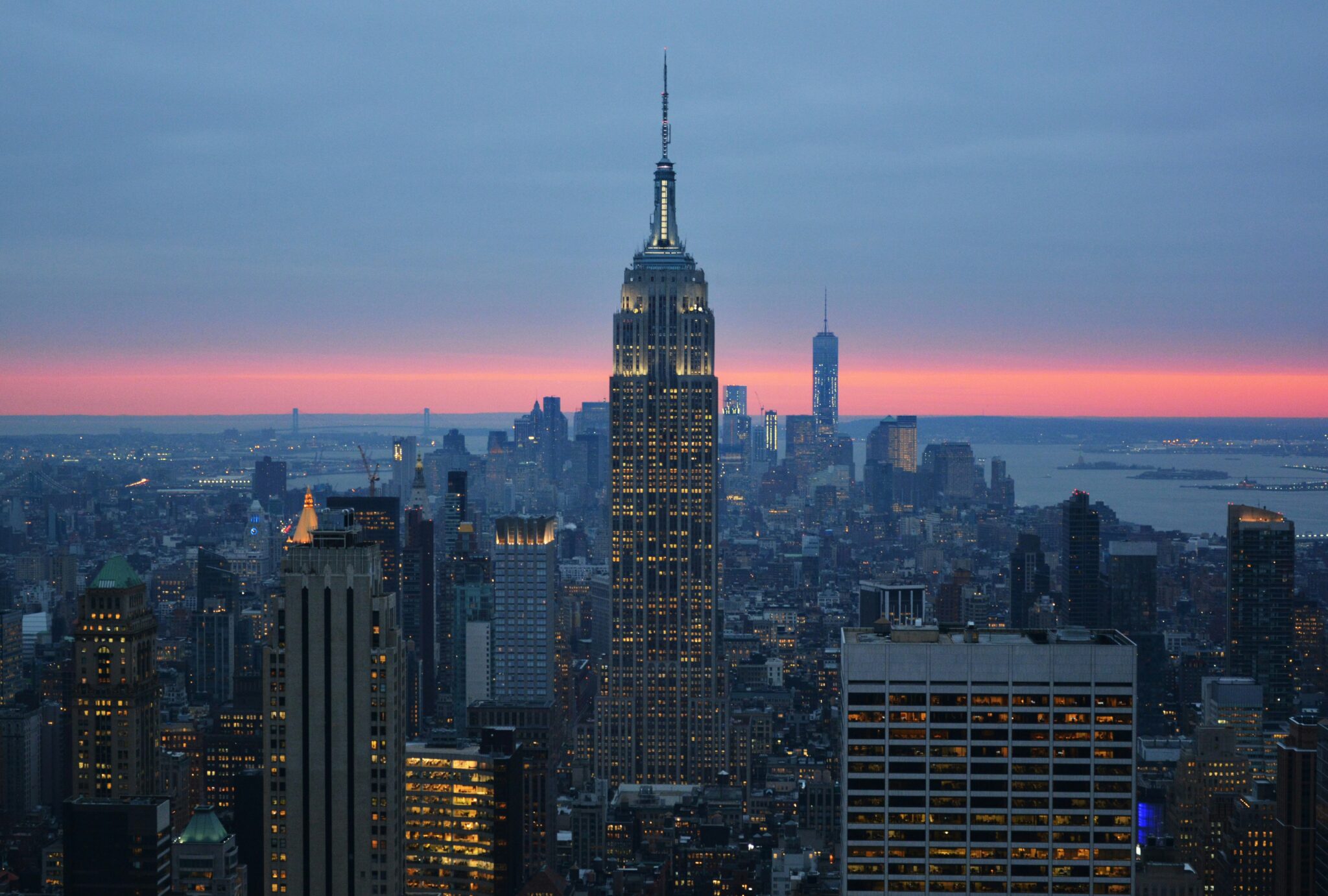 Sunset view of the New York City skyline, featuring the illuminated Empire State Building in the foreground, with the One World Trade Center visible in the background against a pink and blue sky.